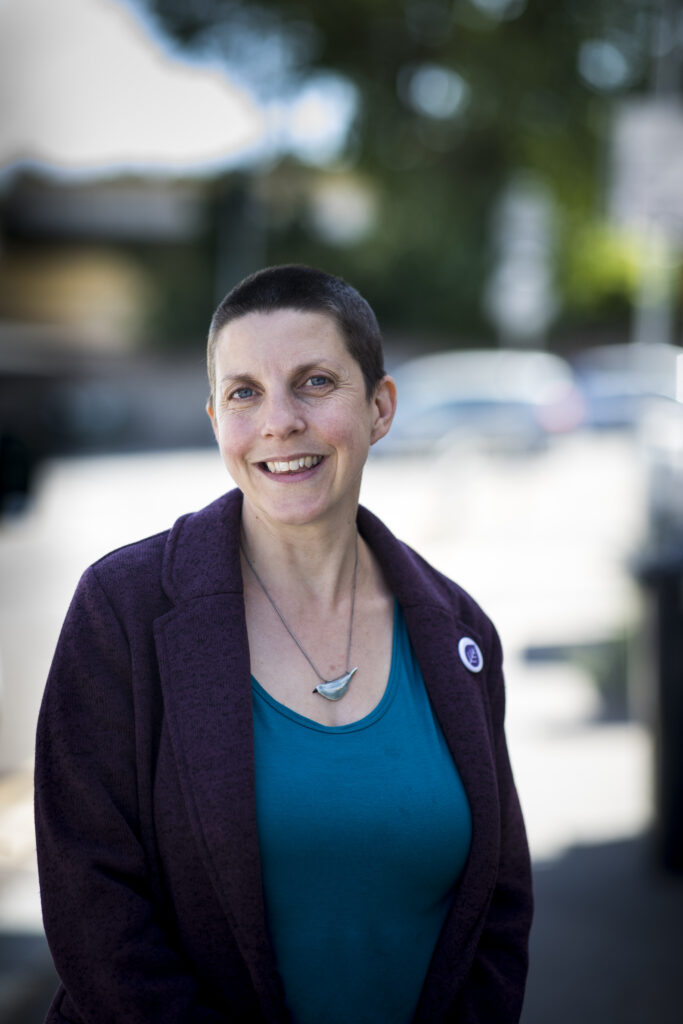 Photo of Jay, the CEO. She is a white woman, has short cropped hair, and is wearing a blue top and a cardigan. She smiles at the camera.