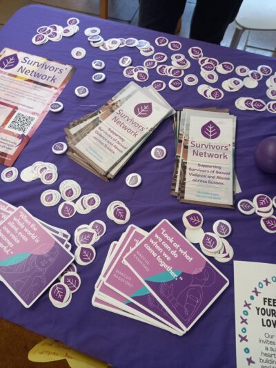 Photo of a Survivors' Network event stall, featuring stickers, leaflets, postcards and posters displayed on a purple tablecloth.