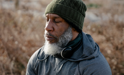 Photo of a self-identifying older man practising mindfulness meditation. The man has light Black skin and grey facial hair. The man is wearing a green hat, grey activewear and black headphones. He is sitting outside surrounded by nature and has his eyes closed.