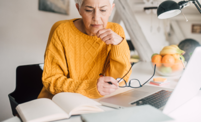 Photo of a self-identifying older woman with white skin and short white hair. The woman is wearing a yellow knitted jumper and is sitting at a desk, holding a pair of black glasses and reading over notes and a laptop.