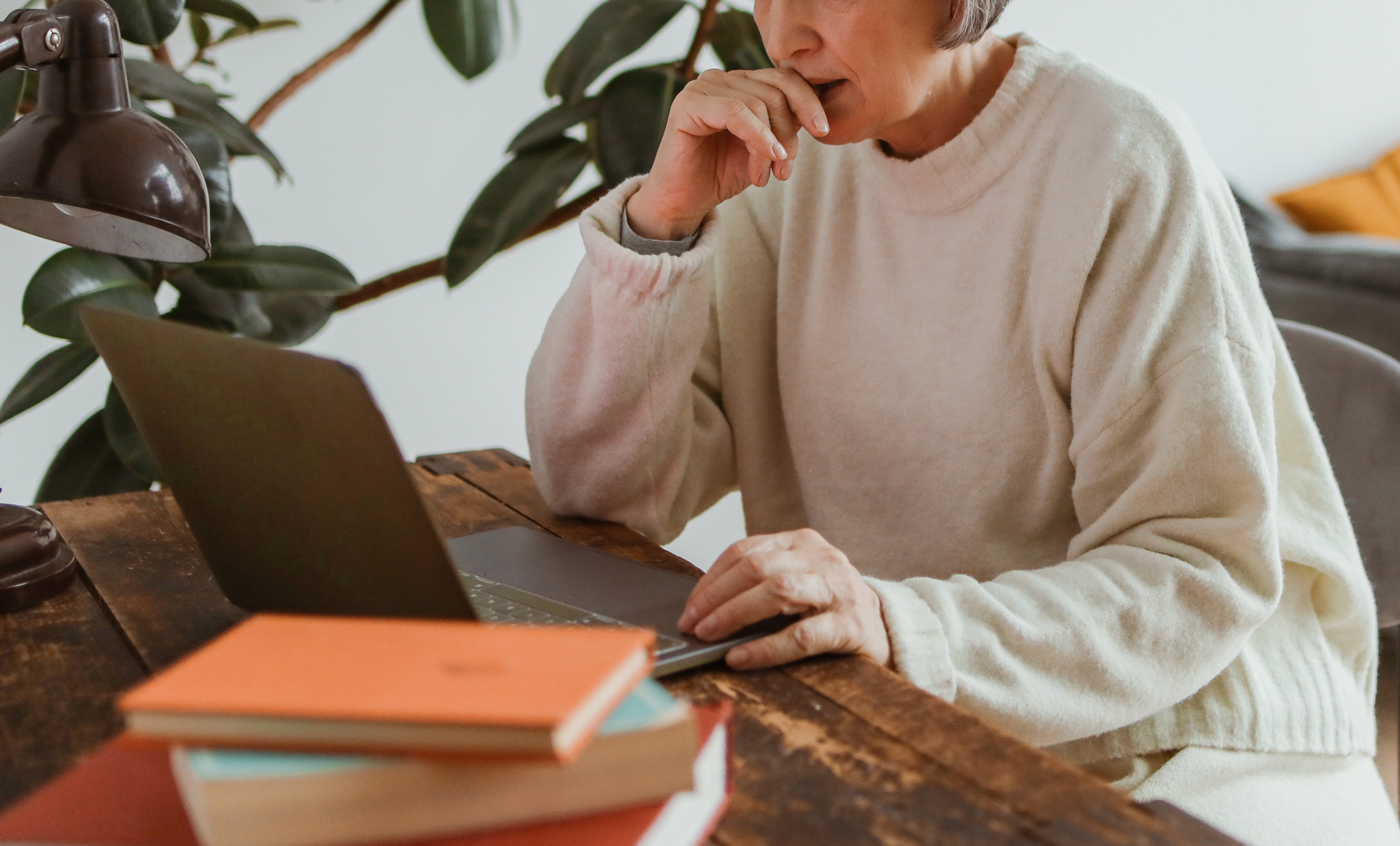 Photo of a self-identifying older woman with white skin and short grey hair. The woman is wearing a cream jumper and sitting at a desk using a laptop.